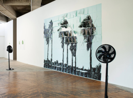 Gran mural de palmeras batidas por el viento frente a dos ventiladores de pie encendidos.